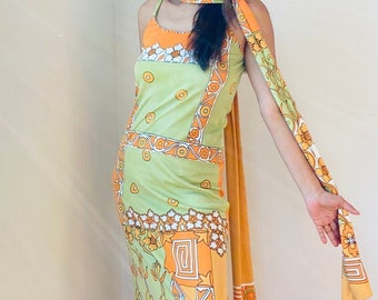 May include: A woman wearing a green and orange patterned dress with a matching orange scarf. The dress has a geometric design with floral accents.