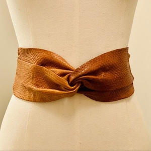 Peut inclure: Une ceinture en cuir marron avec un design noué. La ceinture est large et a une surface texturée.