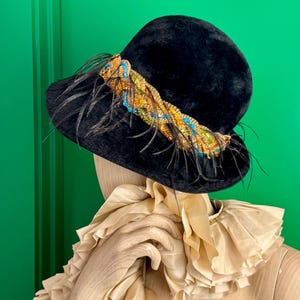 May include: A black hat with a decorative band of colorful beads and feathers. The hat is displayed on a mannequin with a ruffled collar.