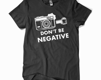 May include: Black t-shirt with a white graphic of a camera and film roll. The text "DON'T BE NEGATIVE" is printed below the camera illustration. The shirt has a crew neck and short sleeves.