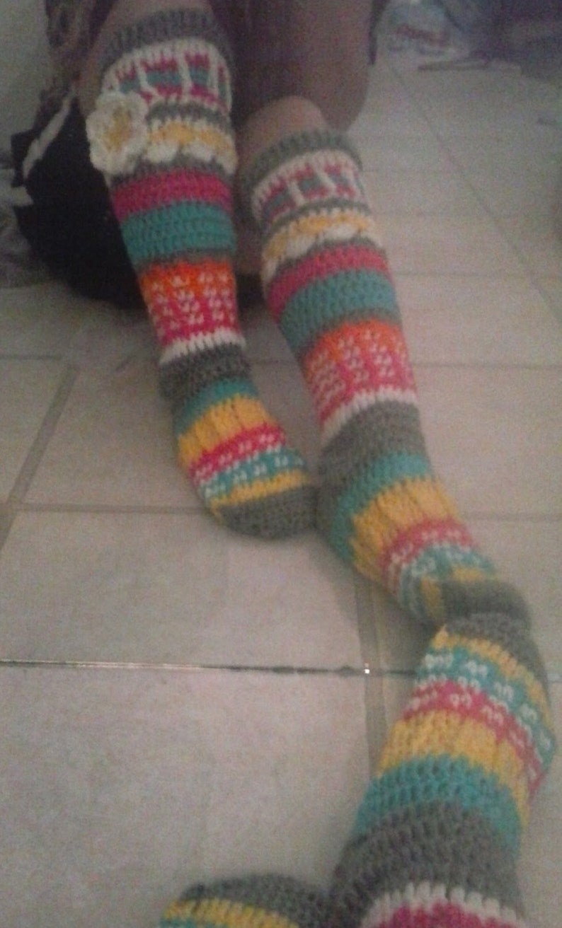 May include: A pair of colorful crocheted leg warmers with a flower embellishment. The leg warmers are made with a variety of colors, including gray, teal, pink, yellow, and white.