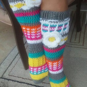 May include: A pair of colorful crochet leg warmers with a white flower design. The leg warmers are made with grey, white, yellow, orange, pink, and turquoise yarn.