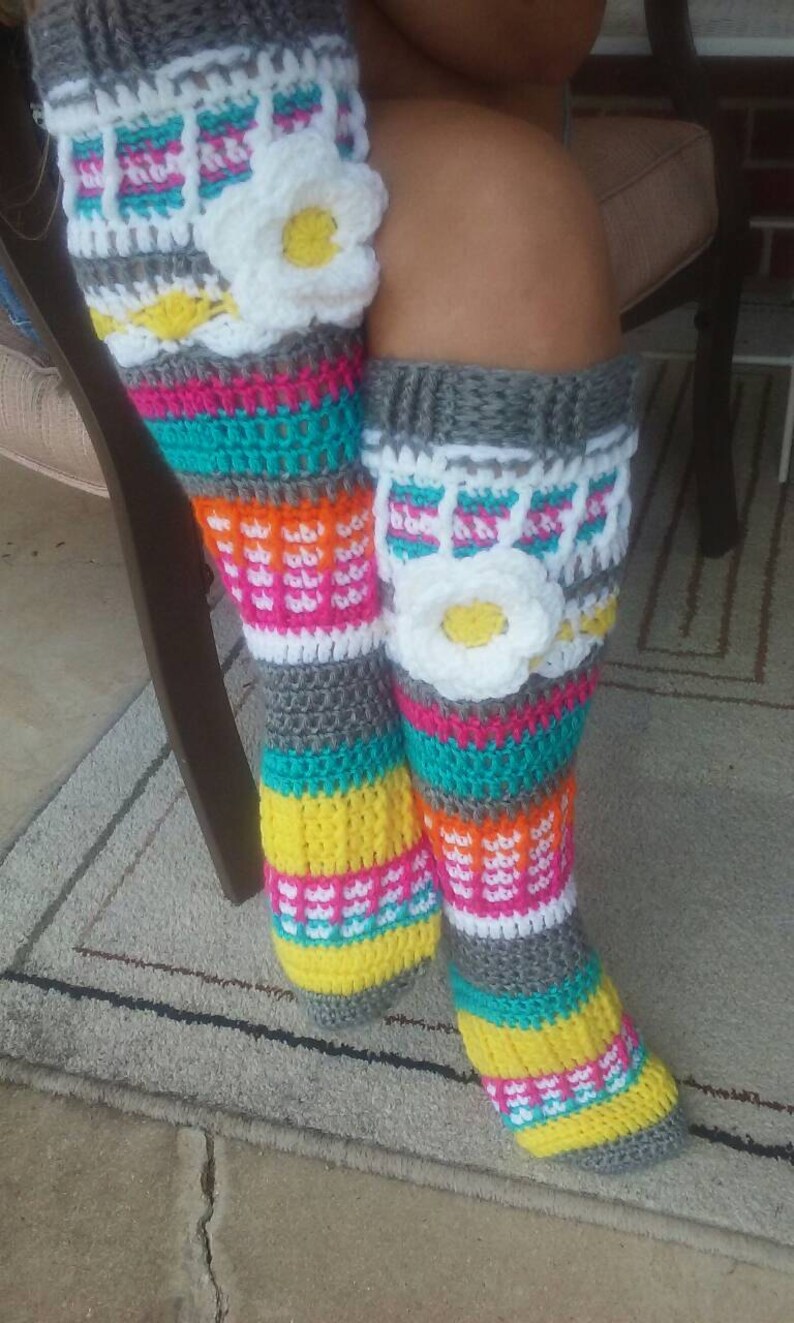 May include: A pair of colorful crochet leg warmers with a white flower design. The leg warmers are made with grey, white, yellow, orange, pink, and turquoise yarn.