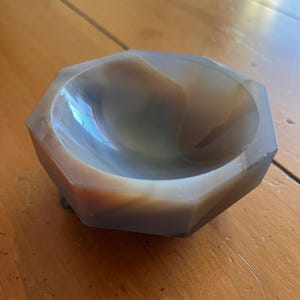 May include: A faceted, agate bowl with a polished interior. The bowl's colors range from light brown to gray, with a translucent quality. The bowl has a geometric, octagonal shape and is likely used for decorative purposes.