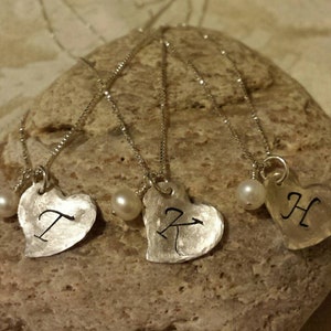 May include: Three silver heart necklaces with initial charms. Each necklace has a small white pearl bead. The initials are T, K, and H.