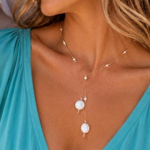 Op de afbeelding: Een gouden ketting met kleine witte parels die over de lengte zijn verdeeld en uitmonden in een Y-vormig ontwerp. Het hangende gedeelte bevat twee grotere, platte, ronde witte parels die op verschillende lengtes aan dunne gouden kettinkjes hangen.