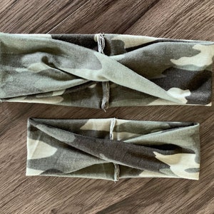 May include: Two camouflage print headbands in shades of green, gray, and black. The headbands are twisted at the front and appear to be made of soft fabric. One headband is smaller than the other.