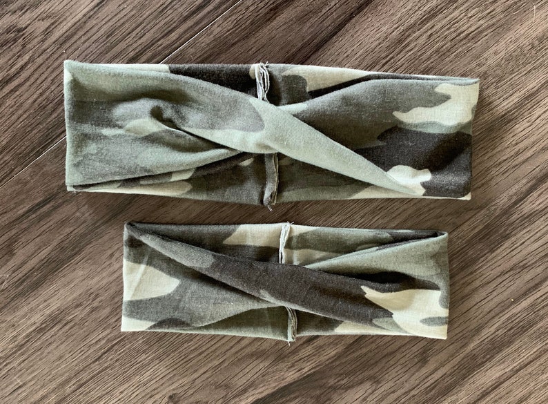 May include: Two camouflage print headbands in shades of green, gray, and black. The headbands are twisted at the front and appear to be made of soft fabric. One headband is smaller than the other.