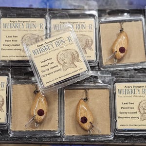 May include: A collection of wooden fishing lures in clear plastic packaging. Each lure is teardrop-shaped with a dark red accent and a hook. The packaging includes the text "WHISKEY RUN-R" and "Angry Sturgeon Lures".