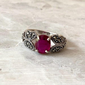 May include: A silver ring featuring an oval, deep red gemstone. The ring's band is adorned with intricate, dark-colored accents, creating a vintage or antique aesthetic. The gemstone is the focal point, set prominently in the center.