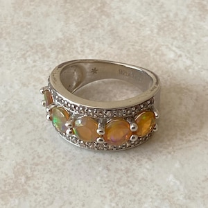May include: Silver ring featuring five oval-cut, orange-toned gemstones. The ring has a band with small, clear accent stones and is marked with "925" and a small star.