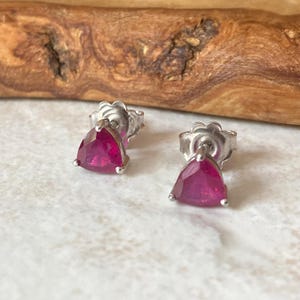 May include: A pair of stud earrings featuring triangular-cut vibrant pink gemstones set in polished silver-toned metal. Each earring has a secure post back and a faceted surface that reflects light, showcasing the deep magenta hue of the stones.