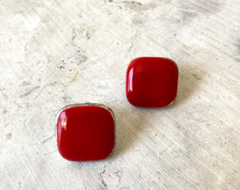 May include: Two square, red earrings with rounded corners and a silver-colored trim. The earrings are set against a light gray, textured background.