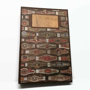 May include: A brown notebook with a cover featuring a collage of vintage cigar band labels. The labels are in various colors and feature different designs, including text, images, and logos. The notebook has a brown spine and a brown cover.