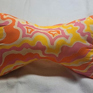 May include: A bone-shaped pillow with a vibrant, retro-style pattern. The fabric features swirling designs in shades of orange, yellow, and pink. The pillow is designed for neck support and has a tie closure.
