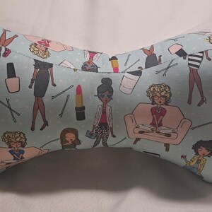 May include: A light blue, bone-shaped reading pillow with a repeating pattern of fashion illustrations, including women, lipstick, and nail polish. The pillow has a small fabric loop on the side.
