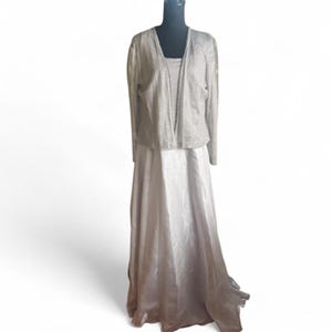 May include: A formal, floor-length dress with a matching jacket. The dress is a light, shimmering color, possibly silver or champagne, with a flowing skirt. The jacket is a similar color and has a textured, dotted pattern. The outfit is displayed on a mannequin.