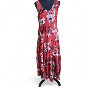 Puede incluir: Vestido largo sin mangas, de color rojo óxido, adornado con un estampado floral en blanco y morado oscuro. El vestido presenta un cuello en V y una falda escalonada, expuesto en un maniquí negro.