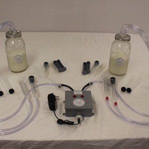 May include: A two-jar milk milking system with a grey control box, clear tubing, and multiple clear graduated cylinders. The system is plugged into an electrical outlet and has two red buttons on the control box.