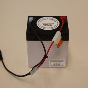 May include: A small, rectangular battery with black and white casing. It has red and black wires connected to it, along with a black cord. The top of the battery has a circular sticker with the text "DANSHA FARMS".
