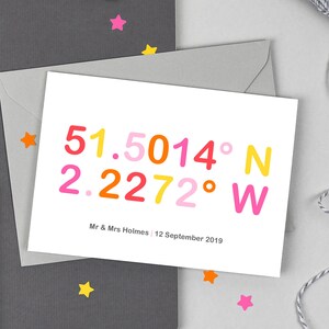 May include: A white card with colourful text that reads "51.5014° N 2.2272° W" and "Mr & Mrs Holmes | 12 September 2019".