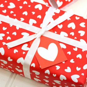 May include: A red gift box with a white ribbon and a white heart on a red tag. The box is wrapped in red paper with a white heart pattern.