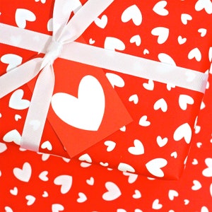 May include: Red gift wrap with a white ribbon and a white heart tag. The gift wrap is covered in a pattern of small white hearts.