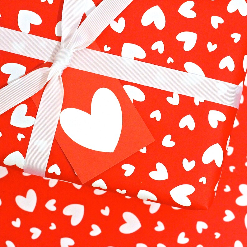 May include: Red gift wrap with a white ribbon and a white heart tag. The gift wrap is covered in a pattern of small white hearts.