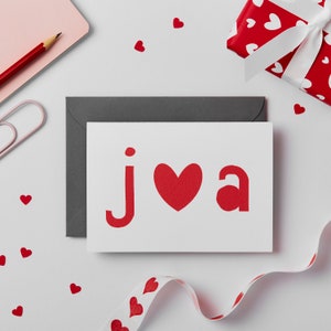 May include: A white card with a red heart and the letters "j" and "a" in red. The card is on a white surface with a gray envelope behind it. There are red hearts scattered around the card.