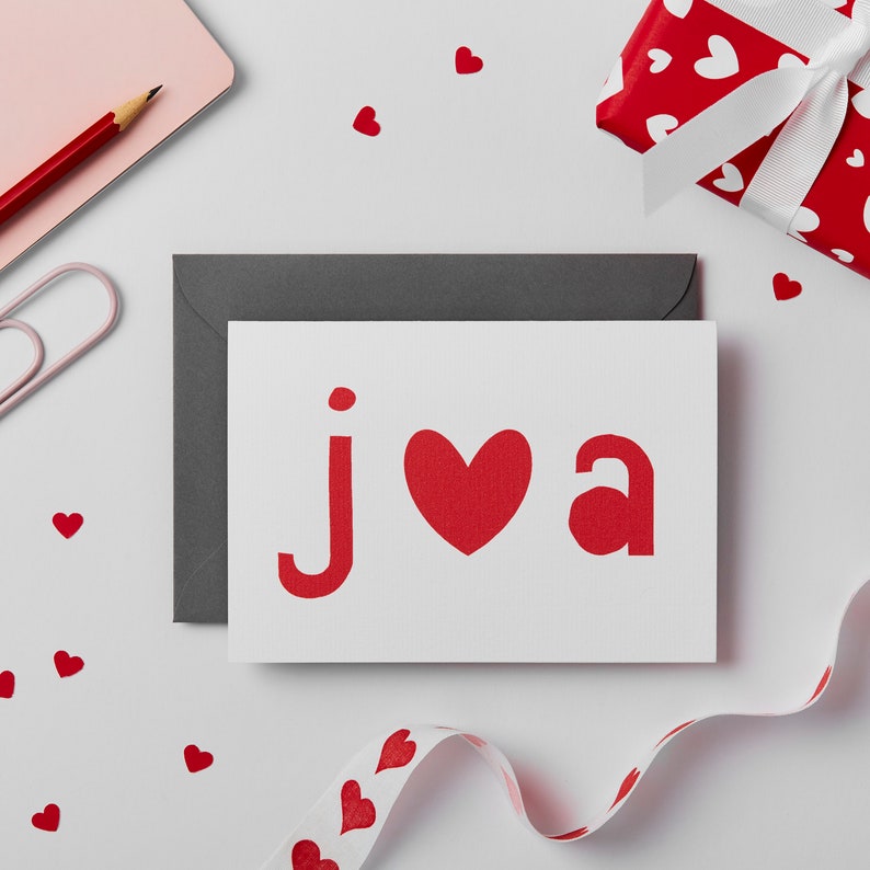 May include: A white card with a red heart and the letters "j" and "a" in red. The card is on a white surface with a gray envelope behind it. There are red hearts scattered around the card.