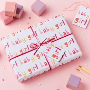 May include: A white gift wrap with a pink, yellow, and red repeating pattern of the words "baby girl". A pink ribbon is tied around the gift wrap with a white gift tag that reads "baby girl".