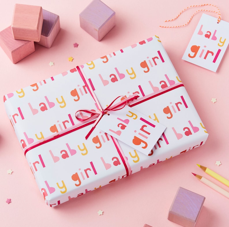 May include: A white gift wrap with a pink, yellow, and red repeating pattern of the words "baby girl". A pink ribbon is tied around the gift wrap with a white gift tag that reads "baby girl".