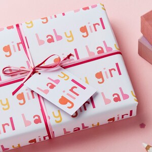 May include: A white gift box wrapped in pink and yellow patterned paper with the words "baby girl" repeated. The box is tied with a pink ribbon and a white gift tag with the words "baby girl" in pink and orange letters.