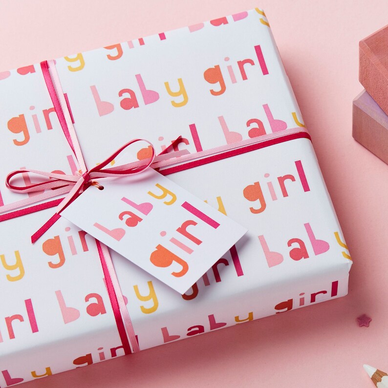 May include: A white gift box wrapped in pink and yellow patterned paper with the words "baby girl" repeated. The box is tied with a pink ribbon and a white gift tag with the words "baby girl" in pink and orange letters.