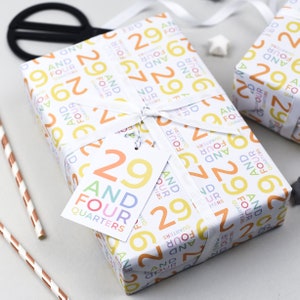 May include: A white gift box wrapped in colorful wrapping paper with a pattern of the numbers 29 and the words "And Four Quarters". The box is tied with a white ribbon and has a white gift tag with the number 29 and the words "And Four Quarters" printed on it.