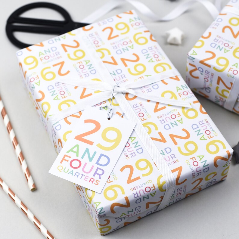 May include: A white gift box wrapped in colorful wrapping paper with a pattern of the numbers 29 and the words "And Four Quarters". The box is tied with a white ribbon and has a white gift tag with the number 29 and the words "And Four Quarters" printed on it.