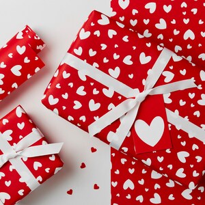May include: Three red gift wrap sheets with white hearts printed on them. The sheets are folded and stacked on top of each other. The top sheet has a white ribbon tied around it.