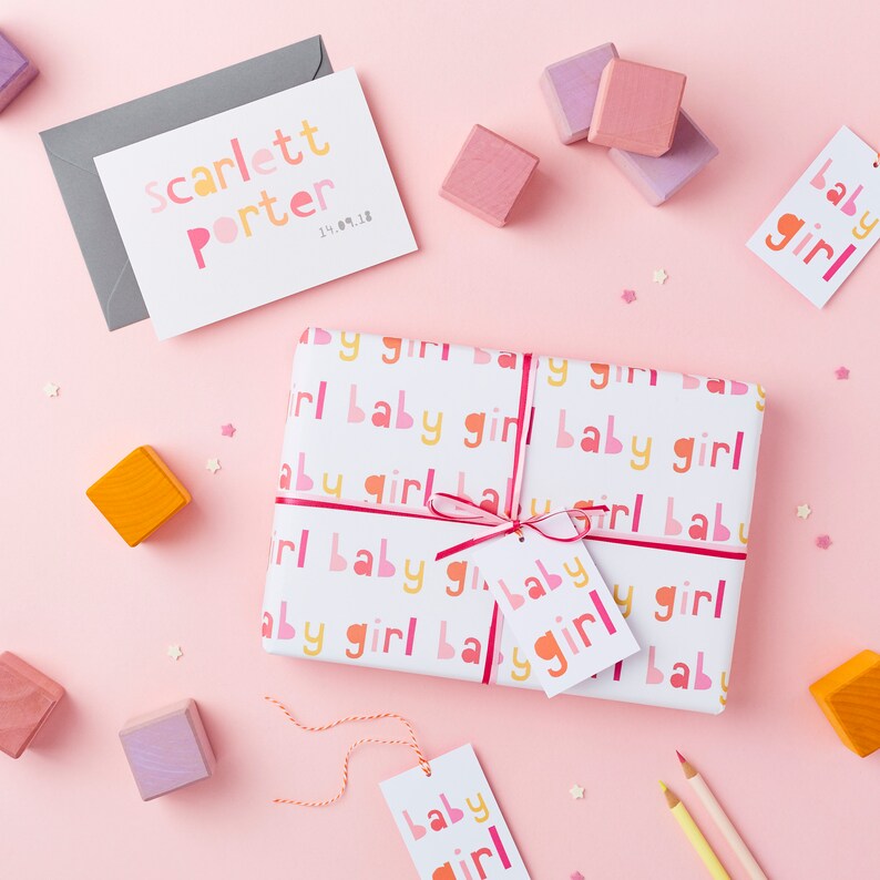 May include: A white gift box wrapped in paper with a pink, orange, and yellow repeating pattern of the words "baby girl". The box is tied with a pink ribbon and has a small white gift tag with the words "baby girl" in pink and orange letters. There are also several small wooden blocks in pink, purple, and orange scattered around the box.