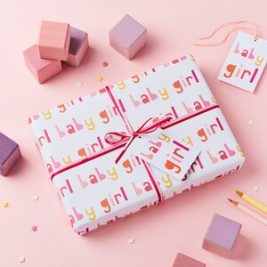 May include: A white gift box wrapped in pink, yellow, and red patterned paper with the words "baby girl" repeated. The box is tied with a pink ribbon and a pink gift tag with the words "baby girl" on it. The box is surrounded by small pink, yellow, and white stars.