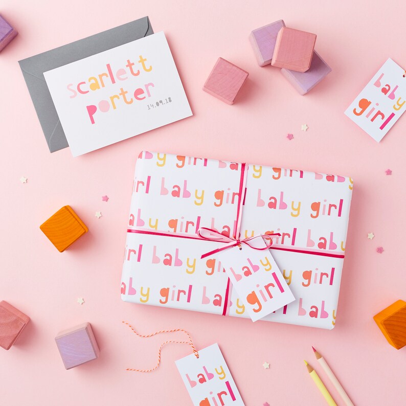 May include: A white gift box wrapped in paper with a pink, orange, and yellow repeating pattern of the words "baby girl". The box is tied with a pink ribbon and has a small white gift tag with the words "baby girl" in pink and orange letters. There are also several small wooden blocks in pink, orange, and purple scattered around the box.