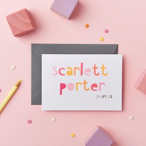 May include: A white card with the name "Scarlett Porter" printed in colorful, block letters. The date "14.09.18" is printed below the name. The card is surrounded by colorful stars and wooden blocks.