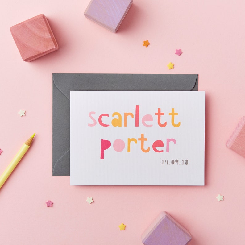 May include: A white card with the name "Scarlett Porter" printed in colorful, block letters. The date "14.09.18" is printed below the name. The card is surrounded by colorful stars and wooden blocks.