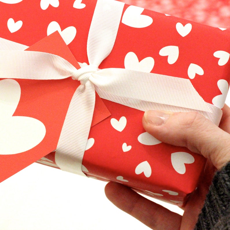 May include: A red gift box with a white ribbon and white hearts printed on the paper. The box is being held by a hand.