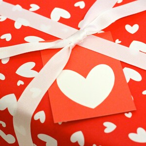 May include: A red gift box with a white ribbon and a white heart on a red tag. The box is wrapped in red paper with white hearts printed on it.