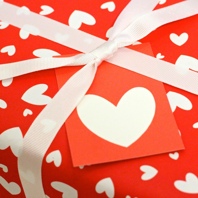 May include: A red gift box with a white ribbon and a white heart on a red tag. The box is wrapped in red paper with white hearts printed on it.