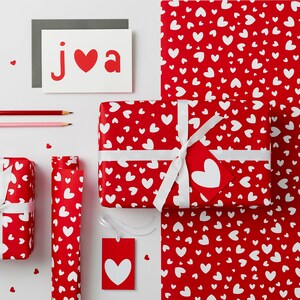 May include: Red and white wrapping paper with a heart pattern. A white ribbon is tied around a gift box wrapped in the paper. A card with the word "JVA" and a red heart is on a white surface.