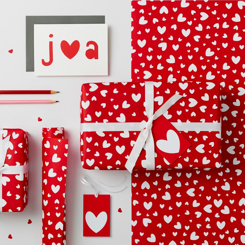 May include: Red and white wrapping paper with a heart pattern. A white ribbon is tied around a gift box wrapped in the paper. A card with the word "JVA" and a red heart is on a white surface.