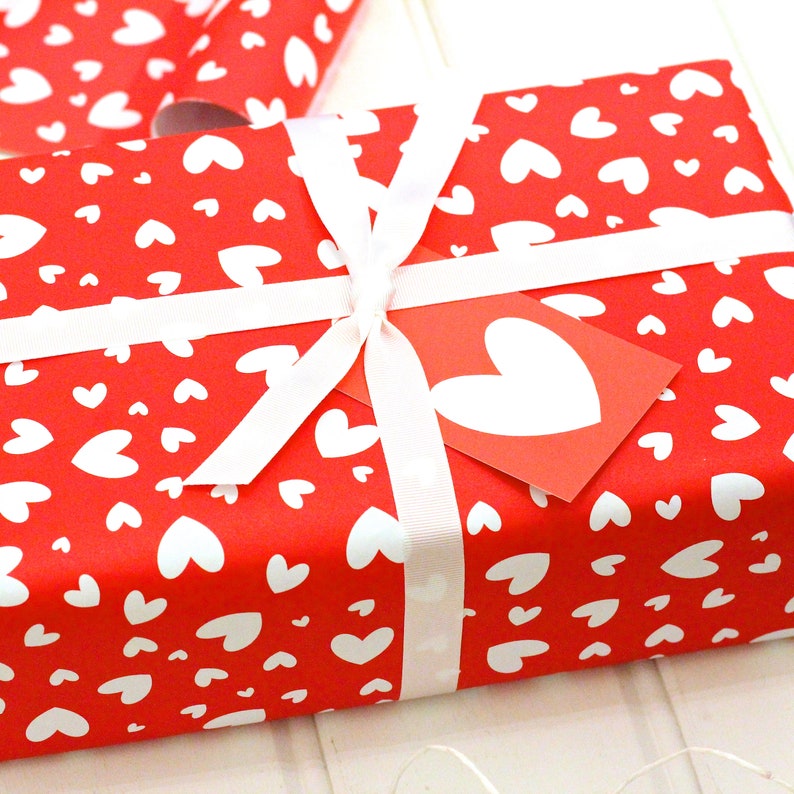 May include: A red gift box with a white ribbon and a white heart tag. The box is wrapped in red paper with a white heart pattern.