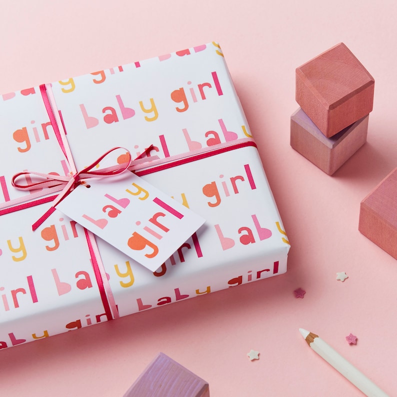 May include: A white gift wrap with a pink ribbon and a gift tag. The gift wrap has a repeating pattern of the words "baby girl" in pink, yellow, and orange. The gift tag is white with the words "baby girl" in pink and orange.