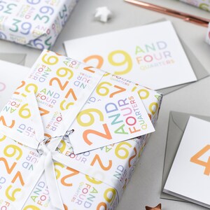 May include: A gift wrapped in white paper with a colorful pattern of numbers and the words "And Four Quarters". The gift is tied with a white ribbon and a tag that says "29 And Four Quarters".